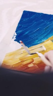 Dikey video. Sanat eseri yaratımı. Kadın ressam. İç mekan resmi. Tanımlanamayan bir kadın, renkli spatula resimleriyle tuvale soyut bir manzara çiziyor..