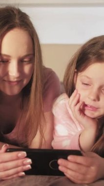 Dikey video. Aile interneti. İnternette sabah. Ev aleti keyfi. Rahatlamış anne kız yatakta telefonda çizgi film tartışmasını izliyor..