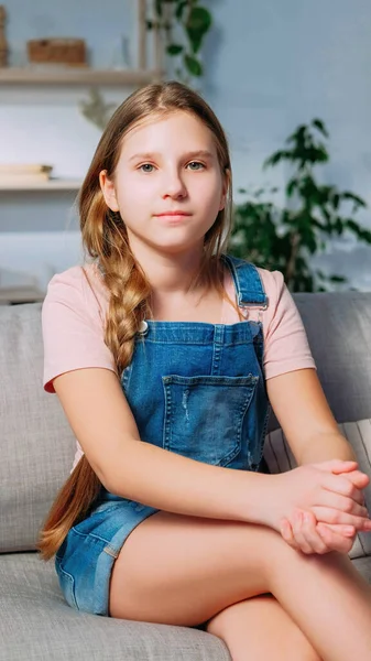 儿童肖像 迷人的孩子 松驰自信的金发姑娘 头戴棉布工作服 头戴辫子 安详的坐在沙发上回家 — 图库照片