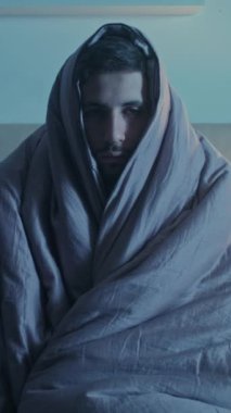 Dikey video. Gece depresyonu. Uyku bozukluğu. Yalnızlık yorgunluğu. Yorgun, uykulu, üzgün, sağlıksız, battaniyeye sarılı, karanlık yatak odasında tek başına uyanık bir adam..