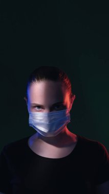 Karantina portresi. Coronavirüs salgını. Virüs korkuyor. Güvenlik önlemleri. Kırmızı mavi ışık koruyucu maskeli kendinden emin kız koyu yeşil fotokopi arkaplanı.