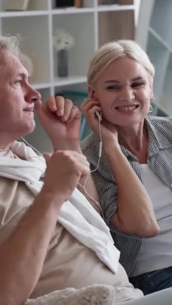 垂直录像 感觉年轻 一对快乐的夫妇喜欢音乐 男人和女人坐在家里的轻便室内 共用耳机听智能手机上的歌曲 — 图库视频影像