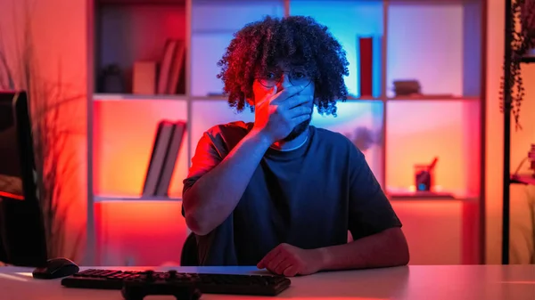 Shocked man. Interesting information. Face expression. Hipster guy covering mouth with hand sitting work desk in dark neon light room interior.