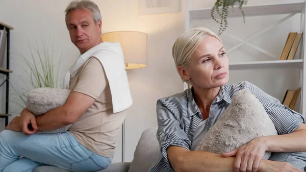 Family quarrel. Offended couple. Disagreement problem. Angry middle-aged man and woman sitting sofa turning back each other in light home interior.