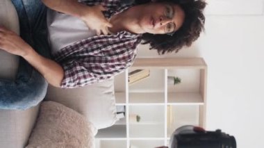 Dikey video. Vlogger yaşam tarzı. Çevrimiçi yayın. Heyecanlı, neşeli, etkileyici adam koltukta konuşuyor. Hafif ev stüdyosunun içinde sosyal medya içeriğini kameraya kaydediyor..