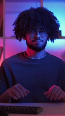 Dikey video. Omuz silkme hareketi. Kafası karışmış bir adam. Meme ifadesi. Karanlık neon ışıklı evin içinde masasında oturan cahil, komik adam el kaldırıyor..