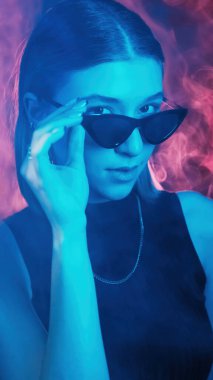 Şık güneş gözlükleri. Renkli ışık portresi. Kendine güvenen neon gözlü kadın koyu mavi kırmızı duman arka planında ciddi bir yüze bakıyor..