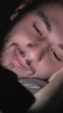 Dikey video. Gece filmi. Gece uykusu. Çevrimiçi eğlence. Rahatlamış adam telefonda komedi programı izliyor. Karanlıkta yatıp gülüyor..