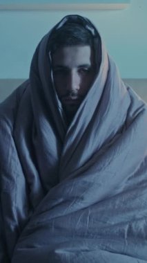 Dikey video. Gece depresyonu. Uyku bozukluğu. Yalnızlık yorgunluğu. Yorgun, uykulu, üzgün, sağlıksız, battaniyeye sarılı, karanlık yatak odasında tek başına uyanık bir adam..