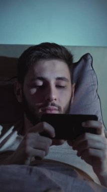 Dikey video. Çevrimiçi film. Geç vakte kadar. İnternet yorgunluğu. Uykucu, yorgun, tembel adam. Akşamları yatakta akıllı telefondan esniyor..