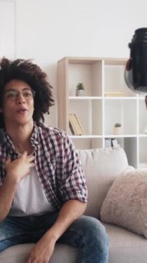 Dikey video. Vlogger yaşam tarzı. Çevrimiçi yayın. Heyecanlı, neşeli, etkileyici adam koltukta konuşuyor. Hafif ev stüdyosunun içinde sosyal medya içeriğini kameraya kaydediyor..