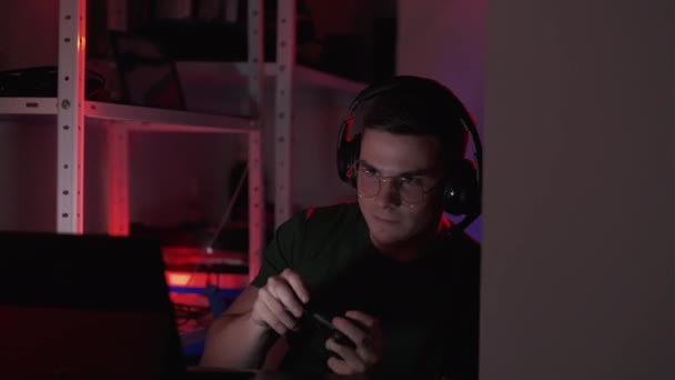 サイバー競争 ビデオゲーム 暗い光の家のインテリアでゲームパッドコンソールを使用してコンピュータ上でオンラインでプレイ眼鏡やヘッドフォンで興奮した男 — ストック動画