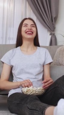 Dikey video. Film izliyordum. Gülen kadın. Evde rahatla. Mutlu kız patlamış mısır yiyor, oturma odasında oturmuş komedi filmi seyrediyor..