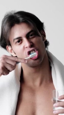 Dikey video. Diş fırçalamak. Ağız bakımı. Omzunda cam su ve diş fırçası olan yakışıklı bir beyaz havlu..