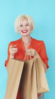 Dikey video. Tatil alışverişi. Mutlu kadın. Şenlik indirimi. Heyecanlı bir kadın mavi poz veren kağıt torbalara bakıyor..