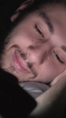 Dikey video. Gece filmi. Gece uykusu. Çevrimiçi eğlence. Rahatlamış adam telefonda komedi programı izliyor. Karanlıkta yatıp gülüyor..