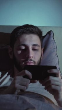 Dikey video. Çevrimiçi film. Geç vakte kadar. İnternet yorgunluğu. Uykucu, yorgun, tembel adam. Akşamları yatakta akıllı telefondan esniyor..