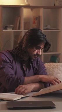 Dikey video. Okuma öğrencisi. Evde öğrenim. Eğitim gelişimi. İşyerinde notlar alan, kitaplarla araştırma yapan konsantre zeki bir adam..