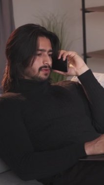 Dikey video. Telefon konuşması. İş görüşmesi. Mobil iletişim. Akıllı telefondan konuşan rahat bir adam evdeki kanepede dizüstü bilgisayar kullanarak çalışıyor..