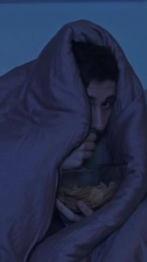 Dikey video. Korku filmi. Ev gerilimi. Korkmuş, rahatsız bir adam battaniyenin altında saklanıyor. TV 'de korku filmi seyrediyor. Mavi ışıklı, karanlık bir odada abur cubur yiyor..