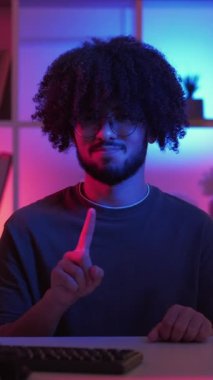 Dikey video. Jest yok. Şüpheci adam. Meme ifadesi. Koyu neon ışıklı evdeki bilgisayarı olan, el sallayan, içi aydınlık bir adam..