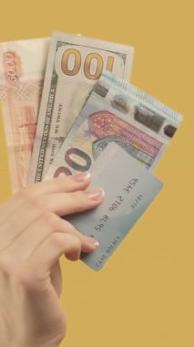 Dikey video. Uluslararası işlem. Yabancı para birimi. Para kredi kartı seti olan sarı arka planda 3 el hareketi yapan bir kadın..
