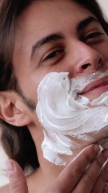 Dikey video. Sabah traşı. Cilt bakımı. Tımar rutini. Çekici, çıplak, yüzü köpüklü yüz bakımı uygulayan pozitif adam..