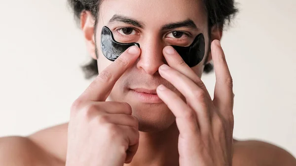 Facial patches. Face treatment. Male beauty care. Attractive guy applying under eye hydrogel black pads isolated on white empty space background.