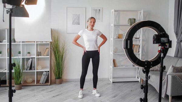 Fitness vlogging. Filming workout. Athletic woman perfect body filming sport training shooting on camera tripod using light equipment exercising in home interior.