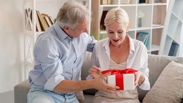 stock image Happy anniversary. Amazed woman. Holiday surprise. Festive middle-aged wife getting pleasant unexpected gift box from husband in light home interior.