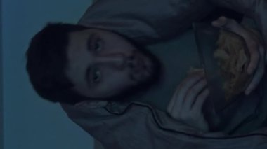 Dikey video. Gece filmi. Ev tatili. Rahatlamış korkusuz adam, battaniyenin altında abur cubur yiyor, TV gerilim filmi izliyor, karanlık yatak odasında..