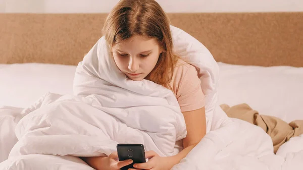 子供中毒 ホームレジャー デジタルライフスタイル 深刻な女の子スクロールフィード上のスマートフォンで午前中にベッドの下で毛布で光の部屋 — ストック写真