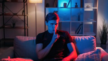 Sır olarak. Sessiz adam. Yüz ifadesi. Komplocu adam parmaklarını ağzının üstüne koydu. Koltuğun üzerinde, neon ışıklı odanın içinde oturuyordu..