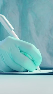 Dikey video. Tıbbi geçmişi. Hasta listesi. Hasta bilgisi. Doktor, mavi koruyucu lastik eldiven ile beyaz masa üzerine kalem ve panoya iş raporu yazdı..