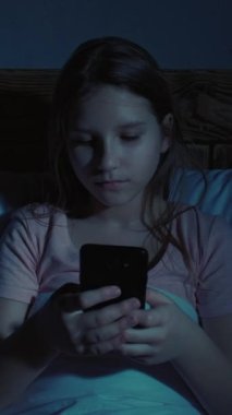 Dikey video. Çocuk uykusuzluğu. Gadget gecesi. İnternet bağımlılığı. Akıllı telefonlu yorgun kız evdeki karanlık odada yatakta mesajlaşıyor..