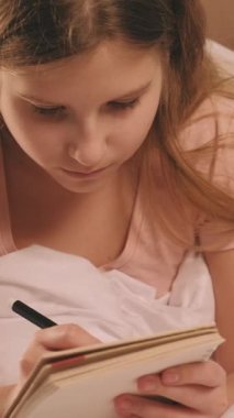 Dikey video. Çocuk günlüğü. Yaratıcı hobi. Çocuk hayal kuruyor. Eskiz defterinden ilham alan kız, sabahları evde rahat beyaz bir battaniyenin içinde dinleniyor..