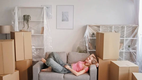 Moving happiness. New home dreaming. Relocation transportation. Relaxed smiling young girl lying on sofa crossed legs resting in white apartment with boxes free space.