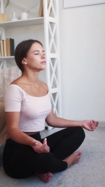 Dikey video. Ev meditasyonu. Farkındalık pratiği. Nilüfer çiçeğinde oturan sakin, genç kadın. Gözleri kapalı, iç mekânda oturuyor..