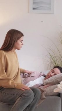 Dikey video. Ev ateşi. Hasta anne. Sevgi dolu kız, sağlıksız bir kadına bakıyor. Yüksek ateş gribi belirtileri, kanepede yatıyor..