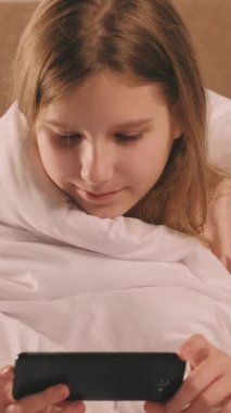 Dikey video. Dijital çocukluk. Çevrimiçi eğlence. Mutlu gülümseyen mutlu kız, sabahları battaniyenin altında komedi programı seyrediyor..