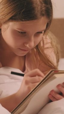 Dikey video. Sabah ödevi. Çocuk eğitimi. Hafta sonu çalışması. Yoğunlaştırılmış odaklanmış ciddi kız battaniyeye uzanıp çizim yapıyor ve ödevini defterde tamamlıyor..