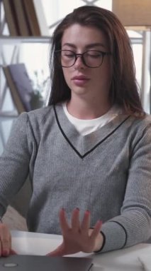 Dikey video. İş zamanı. Yorgun bir kadın. Isınma egzersizleri. Dizüstü bilgisayardaki görevleri bitirdikten sonra boynuna masaj yapan sıradan bir kadın aydınlık odada masa başında oturuyor..