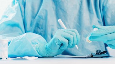 Antibiyotik reçetesi. Tıbbi tavsiye. Doktor mavi koruyucu eldivenler ve ilaç şişesiyle panoya ilaç dozajı yazıyor. Tabletler beyaz masaya su toplamış..