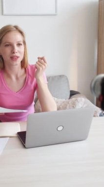 Dikey video. Blog öğretmenliği. Dil hocası. Masada notlar ve dizüstü bilgisayarla gülümseyen kendine güvenen bir kadın, kamera içeriğiyle konuşuyor..