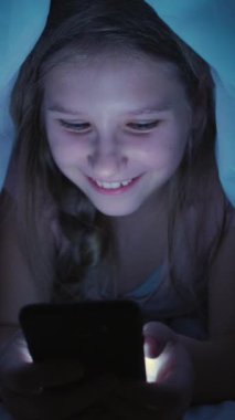 Dikey video. Alfa çocuk eğlencesi. Akıllı telefon gecesi. Heyecanlı mutlu takıntılı kız uyumadan önce battaniyenin altında gülen eğlenceli içeriği izliyor..