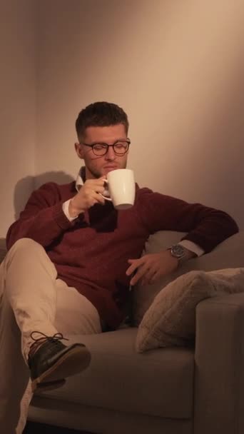 垂直录像 喝咖啡休息 优雅的男人 成功的生活方式松驰的小伙子坐在昏暗的灯光下舒服的沙发上 享受着热饮 — 图库视频影像
