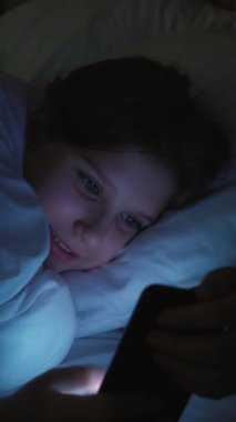 Dikey video. Çocuk bağımlılığı. Dijital bir eğlence. Gece internet sörfü. Mutlu gülümseyen kız sosyal medyada akıllı telefondan besleniyor. Evdeki karanlık odada uzanıyor..