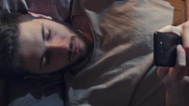 Dikey video. Gadget gecesi. Mobil sohbet. İnternet uykusuzluğu. Sıkılmış yorgun adam mesajlaşan mesajlar kullanıyor. Karanlık yatak odasında geç saatte yatakta yayını kaydırıyor..