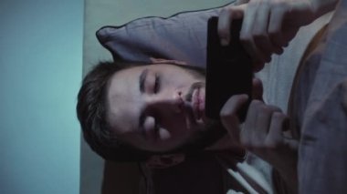 Dikey video. Çevrimiçi film. Geç vakte kadar. İnternet yorgunluğu. Uykucu, yorgun, tembel adam. Akşamları yatakta akıllı telefondan esniyor..