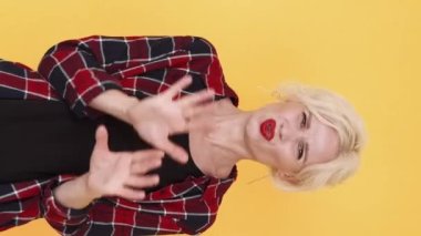 Dikey video. Uzak dur. Reddedilen kadın. Yasak hareket. Yüzünü buruşturan orta yaşlı kadın sarı poz vererek el kol hareketi yapıyor..